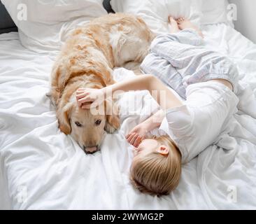 Small Girl giace sul letto giocando con Golden Retriever Dog su biancheria da letto bianca Foto Stock