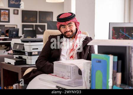 In un elegante ambiente d'ufficio, un gentiluomo arabo siede comodamente sulla sua sedia, trasudando fiducia e calore. Con un sorriso genuino che abbellisce il suo volto, Foto Stock