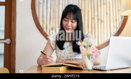 Una bella giovane donna asiatica che si concentra sulla scrittura di qualcosa in un taccuino o sul diario mentre si siede in una caffetteria. le persone e lo stile di vita si fanno beffe Foto Stock