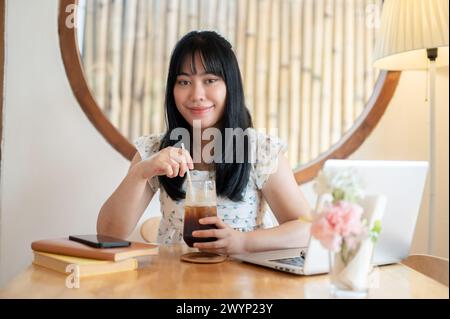 Una giovane donna asiatica bella e allegra, vestita con un bel vestito, si gode il suo caffè ghiacciato mentre si siede a un tavolo in una caffetteria minimalista con Foto Stock