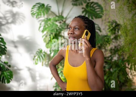 Una ragazza nera interessata fa conversazione con il cellulare circondato da piante d'appartamento tropicali Foto Stock