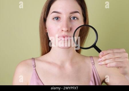 Giovane donna con problemi di acne che tiene la lente d'ingrandimento vicino alla sua pelle su sfondo di oliva Foto Stock