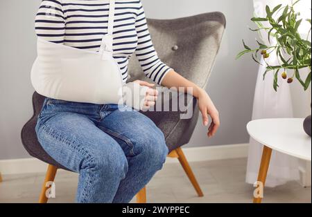 Donna con una ferita al polso che indossa un'imbragatura e una benda seduta su una poltrona a casa Foto Stock