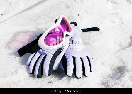 la maschera da sci rosa per bambini con guanti da sci giace su una pista bagnata e innevata in una giornata di sole. Foto Stock