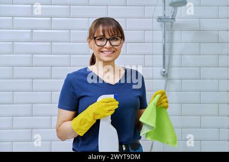 Donna che indossa guanti con un panno detergente spray che esegue la pulizia in bagno Foto Stock