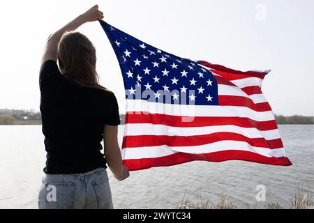 La donna sta con la schiena e tiene una grande bandiera americana al suo fianco. Giornata dell'indipendenza. Simbolo nazionale della libertà degli Stati d'America. Brillante Foto Stock