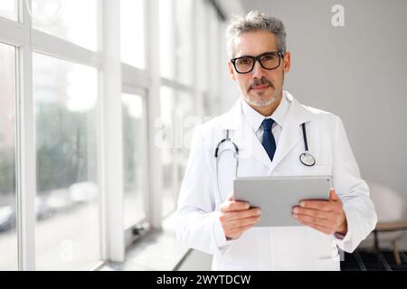 Un medico professionista con i capelli grigi si trova in un ufficio ben illuminato, tiene in mano un tablet digitale e guarda la fotocamera con la massima serietà Foto Stock