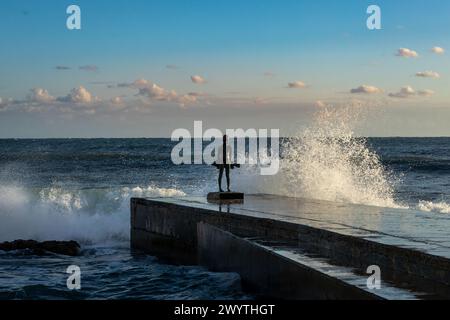 Paphos, Cipro - 23 dicembre 2023: Grandi onde marine che si infrangono sul lungomare di Paphos. Una piccola scultura in bronzo Fisherman sul molo. Foto Stock