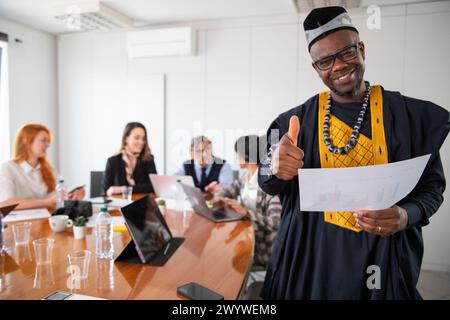 Un uomo d'affari africano durante una riunione si presenta pollice in su, i suoi colleghi lavorano dietro. Foto Stock