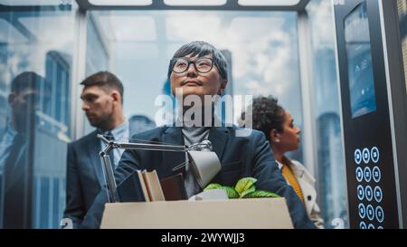 Nuovi dipendenti in arrivo per il primo giorno in ufficio, con ascensore in vetro con diversi colleghi nel moderno business center. Felice manager giapponese con scatola di cartone e accessori per ufficio. Foto Stock