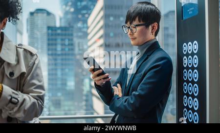 Elegante uomo d'affari giapponese di successo, con ascensore di vetro per l'ufficio nel moderno centro affari. Bell'uomo sorridere mentre si utilizza lo smartphone, scrivere un messaggio di testo, controllare i social media in un ascensore. Foto Stock