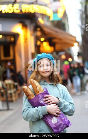 Ritratto di una giovane adolescente francese in berretto blu che tiene le baguette sullo sfondo delle luci serali di un caffè o di un panificio su un narr Foto Stock