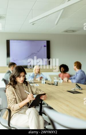 Donna d'affari concentrata con un blocco appunti si stupisce durante una riunione di gruppo, con colleghi impegnati in discussioni in background Foto Stock