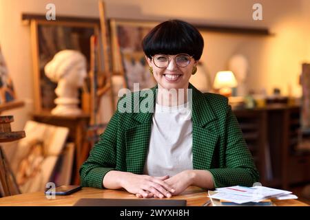 Vista frontale ritratto POV di una donna d'affari sorridente che parla con un computer portatile mentre si siede alla scrivania nello studio d'arte e guarda la fotocamera Foto Stock