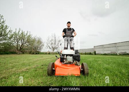 Il giardiniere professionista in abbigliamento protettivo taglia prati verdi utilizzando un moderno rasaerba a benzina senza fili nel cortile posteriore. Giardino stagionale desi Foto Stock