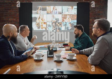 Quattro uomini d'affari, team leader seduti in una sala conferenze, impegnati in una videochiamata con i dipendenti, discutendo delle strategie aziendali Foto Stock