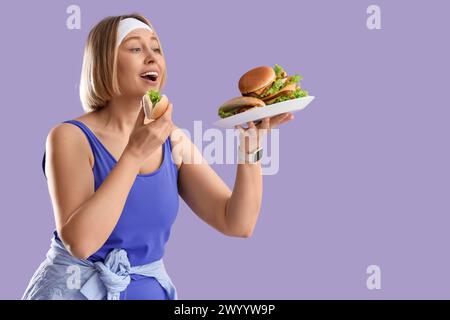 Bella donna sportiva con cibo malsano su sfondo lilla. Concetto di eccesso di cibo Foto Stock