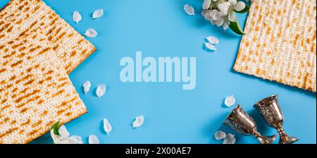 Felice flatlay di passaggio. Calice in metallo, fiori primaverili e matzah tradizionale su sfondo blu. Simboli della festa ebraica. Foto Stock