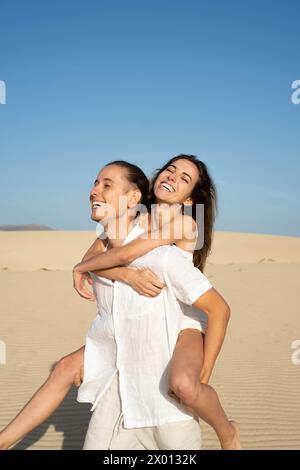 Vista laterale di una coppia allegra che guarda lontano mentre cammina un maschio che porta una femmina a piedi nudi con un sorriso di toothy alla luce del giorno contro una blu senza nuvole Foto Stock