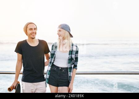 Coppia, skateboard e sorridi sull'oceano per divertirti con spazio per il mockup, avventura e appuntamento sul lungomare. Skater Gen z, persone e ritratti sul lungomare per Foto Stock