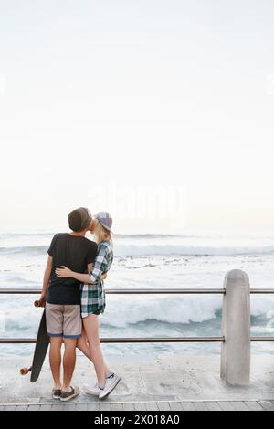 Coppia, baci e romanticismo sulla spiaggia per un appuntamento con spazio per il mockup, avventura e skateboard sul lungomare. Skater, persone e abbracciare la passerella per Foto Stock