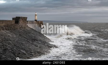 Mari e onde scoscese che si infrangono su un muro di mare e un faro (Porthcawl, Galles del Sud, Regno Unito) in un giorno nuvoloso d'inverno Foto Stock