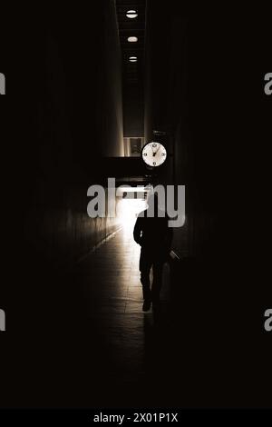Un uomo cammina lungo un corridoio buio con un orologio sul muro. L'immagine ha uno stato d'animo di solitudine e solitudine Foto Stock