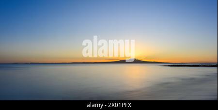 Bagliore dorato sulla cresta dell'isola di Rangitoto mentre il sole sorge sopra l'orizzonte. Milford Beach. Auckland. Foto Stock