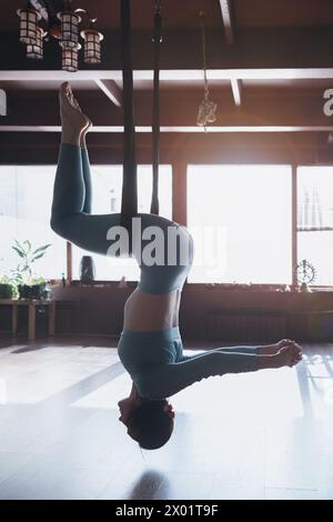 Donna che fa yoga aereo, esegue l'esercizio Utkatasana, sedia invertita in posa su un'amaca, si allena da sola in abbigliamento sportivo in uno studio di yoga Foto Stock
