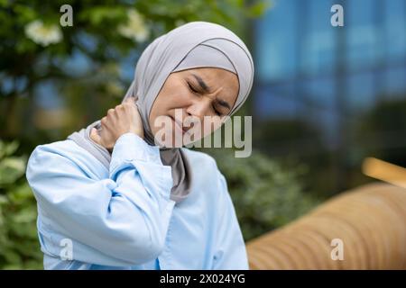 Ritratto di donna araba malsana che piega la testa con gli occhi chiusi mentre sfrega il collo su sfondo sfocato. Donna malata che ha infiammazione nei muscoli e massaggia la spalla come terapia quotidiana all'aperto. Foto Stock