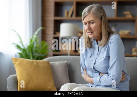 Donna anziana che tiene lo stomaco con entrambe le braccia mentre soffre di spasmi gravi e dolori addominali al chiuso. Donna scioccante con problemi di salute avendo crampi nel sistema digestivo durante il giorno. Foto Stock