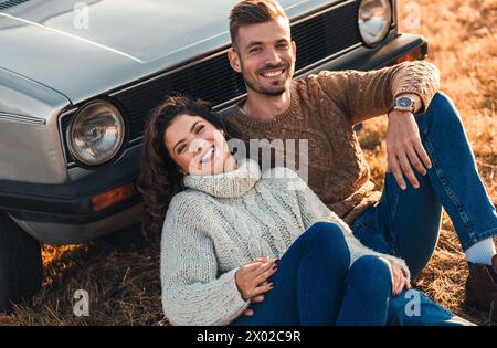 Bella giovane coppia che si diverte insieme all'aperto seduto su un prato appoggiato su un'auto vecchia che si abbraccia a vicenda. Foto Stock