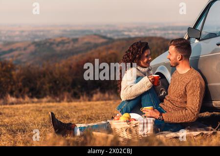 Bella coppia giovane che si gode un picnic al tramonto. Bevono tè e si siedono in un prato appoggiato a un'auto vecchio stile. Foto Stock