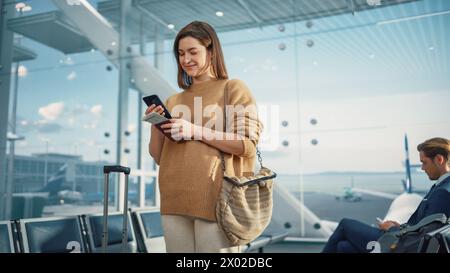 Terminal dell'aeroporto: Donna caucasica felice di viaggiare in attesa ai cancelli di volo per l'imbarco in aereo, utilizza smartphone mobile, controlla la destinazione del viaggio su Internet. Donna bianca sorridente in vacanza Foto Stock
