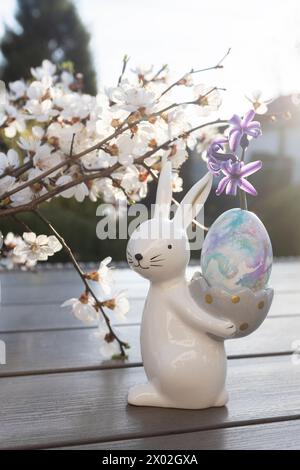 Il coniglietto pasquale in porcellana souvenir regge un uovo sullo sfondo dei rami primaverili di alberi bianchi in fiore. Arredamento per le vacanze Foto Stock