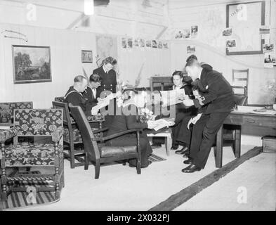 AL "SAILOR'S REST" DI HARWICH. 27 LUGLIO 1944, HARWICH. È STATA APERTA UNA VECCHIA STALLA CONVERTITA DA RATING IN UNA SALA DI RIPOSO E INFORMAZIONE. - L'ufficiale di collegamento per i servizi del capo che gestisce la sala relax che chiacchiera con i marinai mentre si rilassano su poltrone Foto Stock