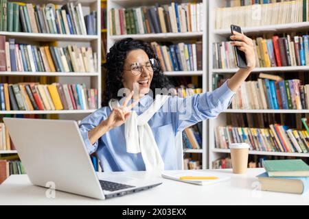 Allegra studentessa ispanica scatta un selfie giocoso tra i libri in una biblioteca, esemplificando la vita e la felicità delle studentesse. Foto Stock