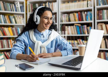 Uno studente concentrato si impegna a seguire l'apprendimento online in una libreria, utilizzando le cuffie e prendendo appunti accanto a una tazza di caffè. Foto Stock