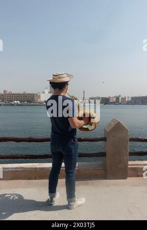 Giovane musicista che suona il corno francese sulla riva del torrente nel quartiere storico di al Shindagha, Dubai, Emirati Arabi Uniti. Foto Stock