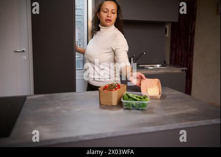 Bella donna, casalinga che prende una scatola di cartone con formaggio dal frigorifero, la mette sul tavolo della cucina con gli ingredienti, preparando guarigione Foto Stock