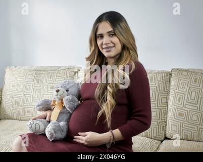 Ragazza latina incinta sorridente alla telecamera, che tiene in mano un orsacchiotto per la sua futura figlia. Foto Stock