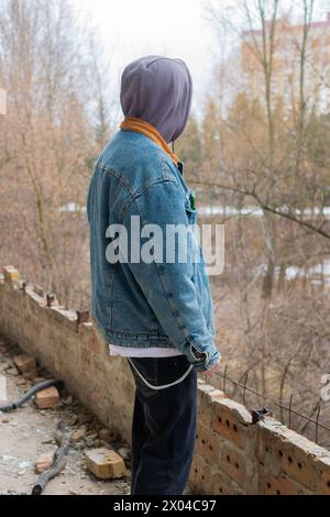 Un bel ragazzo con una giacca in denim e un cappuccio grigio si posa sullo sfondo di un edificio distrutto e abbandonato e di alberi secchi senza foglie. Foto Stock