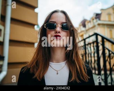 Una donna con gli occhiali da sole, una camicia bianca e una giacca nera si staglia tranquillamente sulla strada. Foto Stock