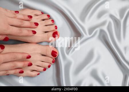 Donna che mostra eleganti unghie del piede dopo la procedura di pedicure e mani curate con lucidatura rossa su tessuto di seta grigio, vista dall'alto. Spazio per il testo Foto Stock