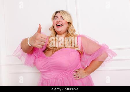 Modello femminile entusiasta e curvato che indossa abiti eleganti con la mano in vita che mostra il pollice in alto e in piedi su sfondo bianco in studio Foto Stock