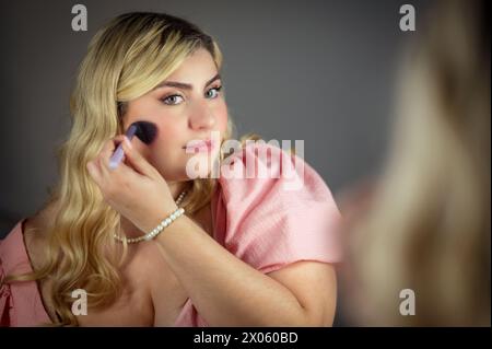 Donna sicura di sé in un abito elegante che guarda lo specchio mentre utilizza la spazzola per il trucco per applicare il cosmetico Foto Stock