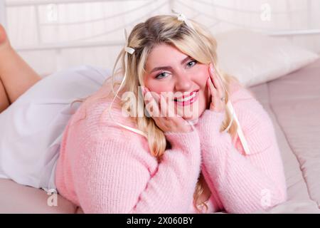 Una giovane donna sorridente e di grandi dimensioni che indossa un abbigliamento casual e si rilassa su un comodo letto con le mani sul mento e guarda la macchina fotografica in una stanza luminosa Foto Stock