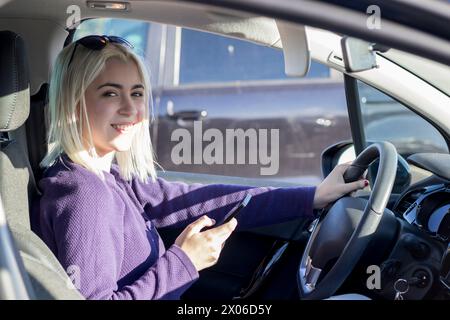 Allegra donna bionda in un maglione viola all'interno di un'auto, che tiene uno smartphone con una mano mentre l'altra è al volante, indossando la Sunglasse Foto Stock
