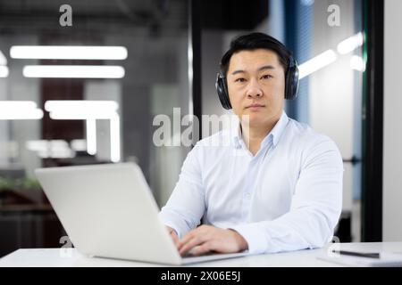 Uomo d'affari asiatico che lavora al suo notebook indossando le cuffie in un ufficio moderno. Foto Stock