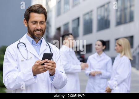Un giovane medico sorridente si trova fuori dalla clinica con un cappotto bianco e usa il telefono. dietro, un gruppo interrazziale di colleghi parla tra loro. Foto Stock